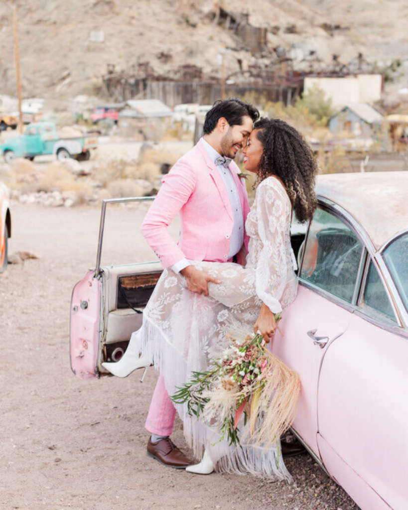 couple kissing leaning up against a pink cadillac with a desert ghost town background
