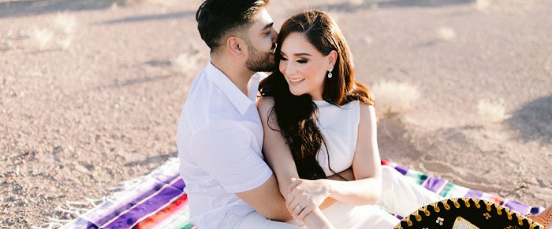 Groom kissing the bride on the side of her head while sitting on a colorful mexican blanket with a sombrero peeking into the shot