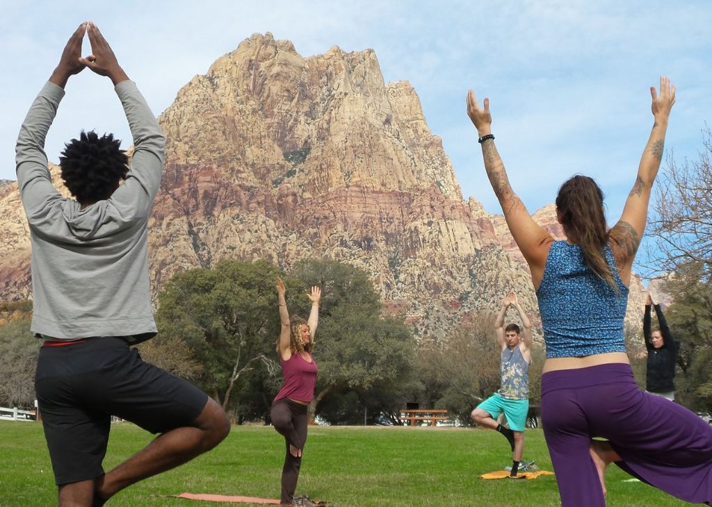 Four people doing morning yoga on green grass with blue sky and tan, red mountain as backdrop