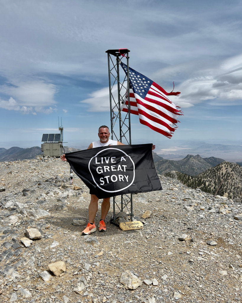 Trent holding a flag that says "Live a Great Story" in front of the American flag on the Peak of Mount Charleston. It's a blue cloudy sky day.