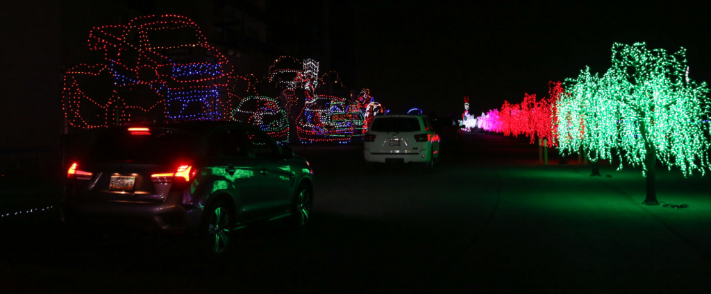 Vehicles driving through monster truck light displays on one side and red, green, white and pink trees on the other at Glittering Lights at Las Vegas Motor Speedway