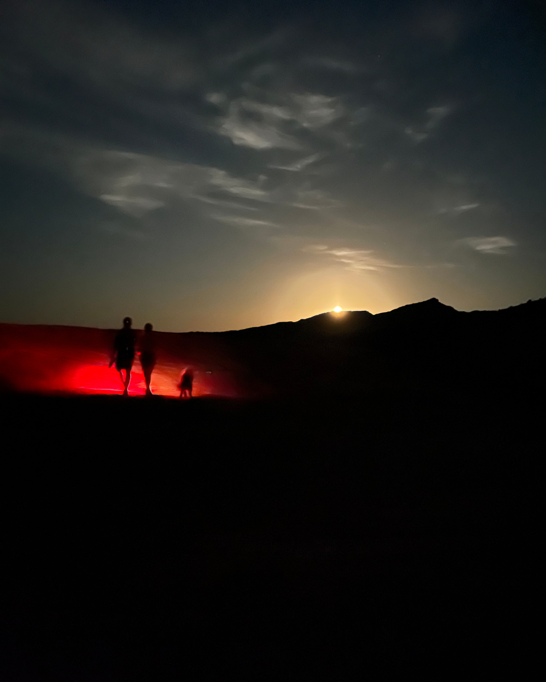 A moonlit night as the moon crests over the Fire Wave Trail with clouds in the sky and three people in the distance walking with their red lights.