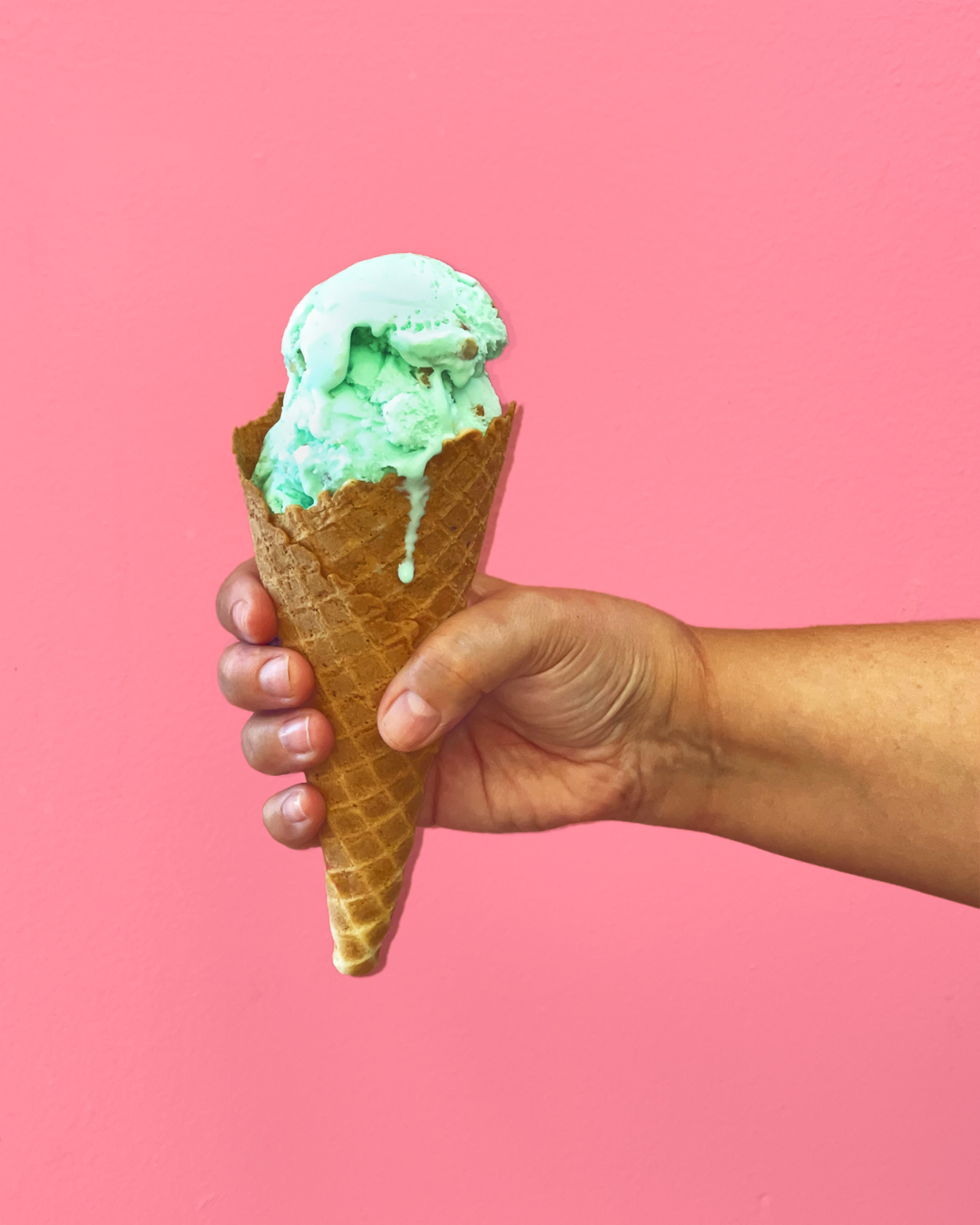 A bubblegum pink background and a hand holding a waffle cone with a scoop of mint ice cream.