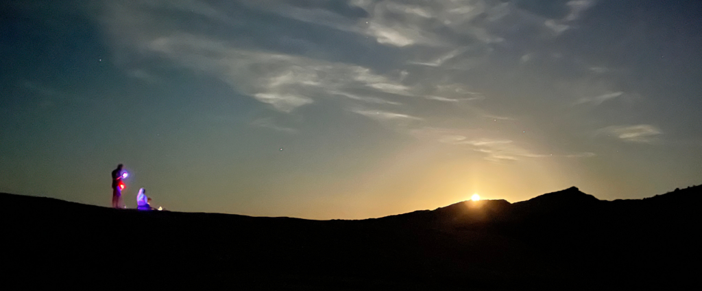 Two people on the Fire Wave Trail Rocks watching the moon crest over the Valley of Fire.