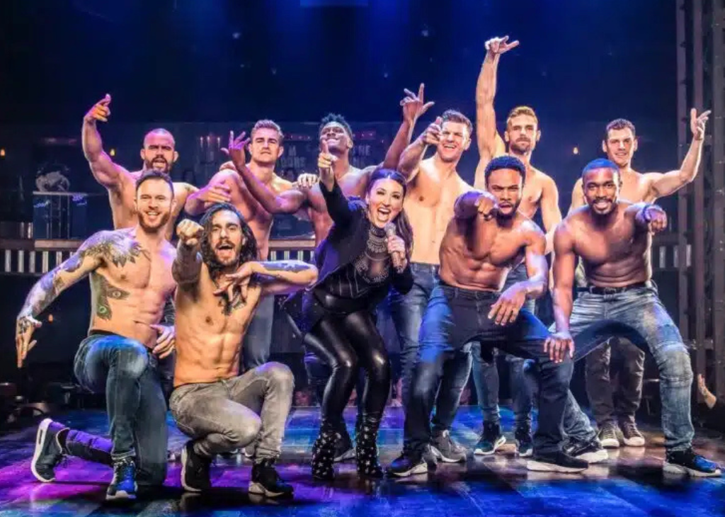 A girl in black leather in the midst of the magic mike entertainers on stage with jeans and no shirts some fist pumping