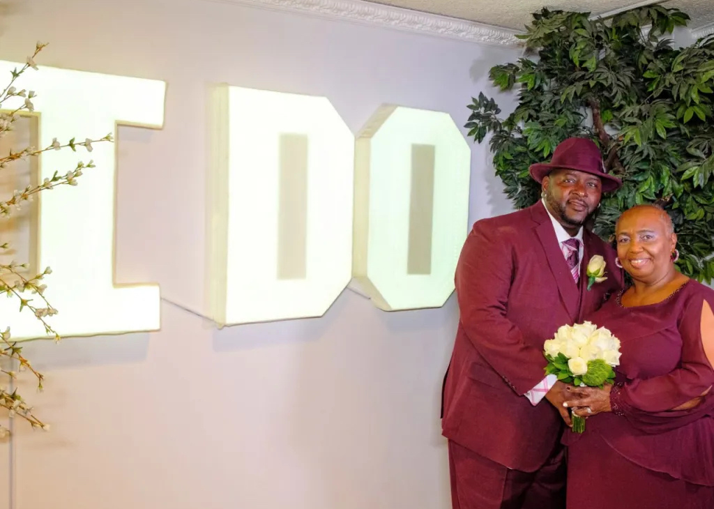 A couple in Maroon formal wear standing in front of a 3-D sign on the wall that says I DO