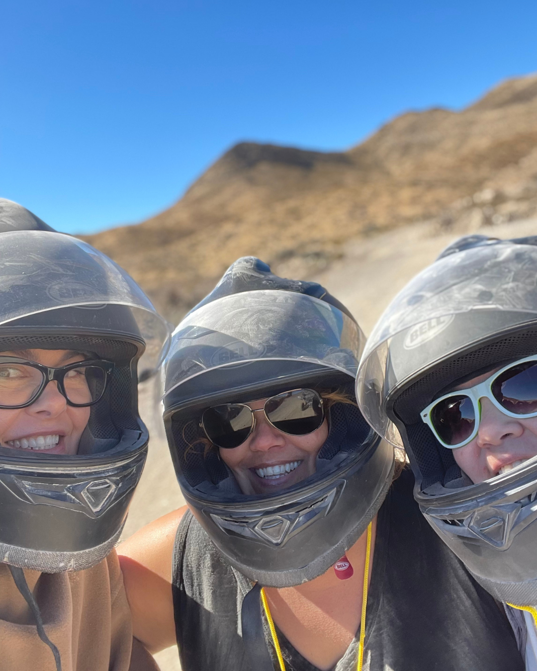 Three girls with dusty UTV helmets on and a blue sky and brown mountain background.