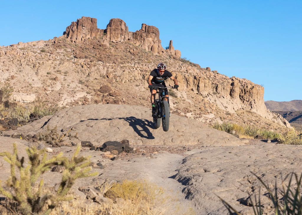 A man on a electric bike jumping from a hill along a trail with desert rock formations the background.