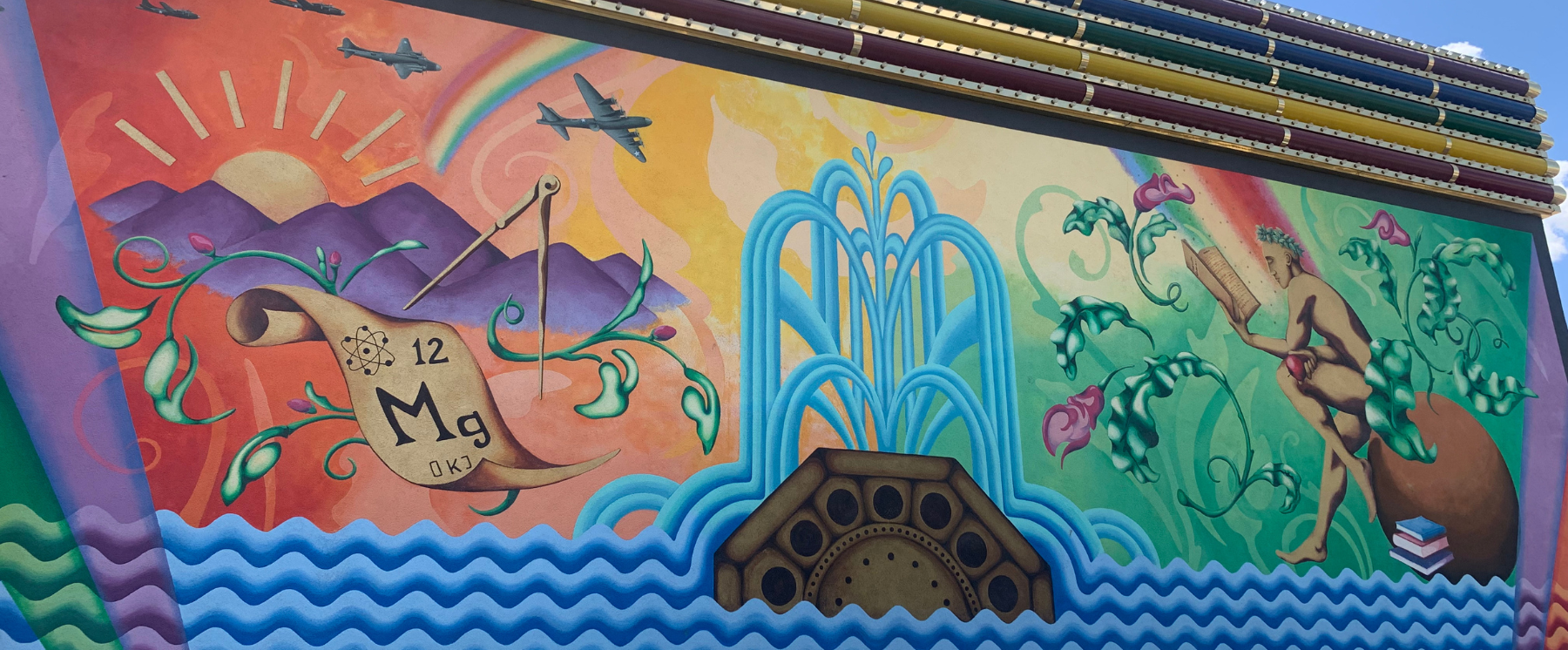 A Mural on the Rainbow Club parking lot wall showing water, rainbows and mermaid reading a book on a rock with mountains and beach.