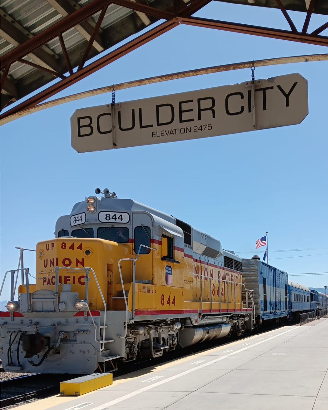 A yellow Union Pacific train engine in the depot with a Boulder City sign on clear blue sky day.