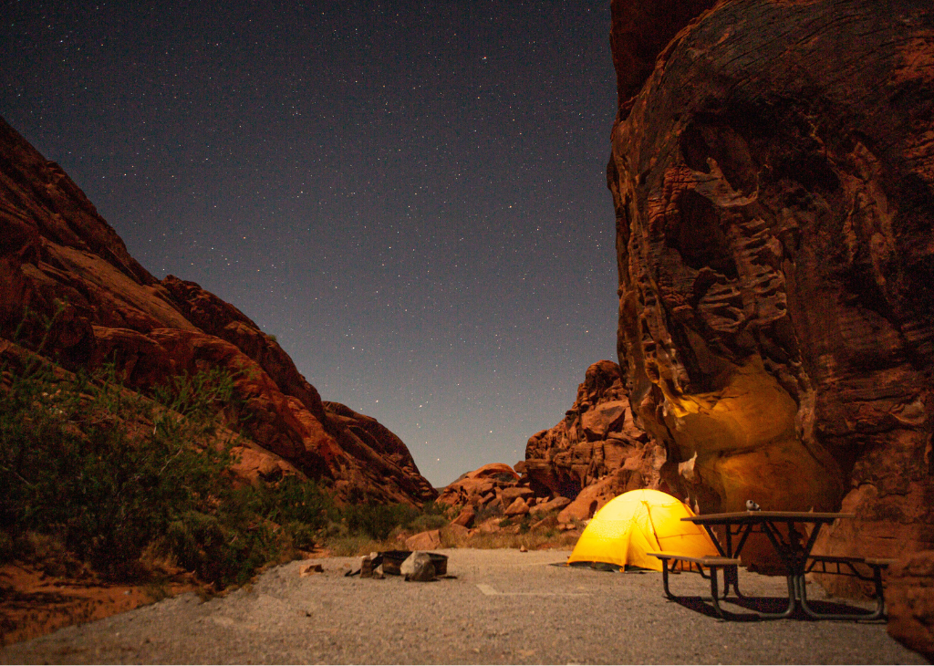 What's New Fall 2023 - this pic shows a tent in yellow and orange glow in between two giant red rocks and sandy path with a starry night above at Valley of Fire.