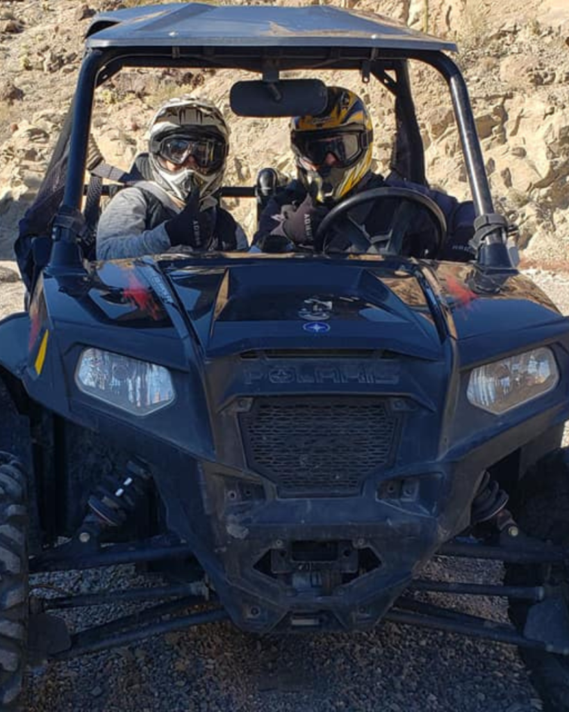 two people with helmets in a black/blue Polaris with the desert terrain behind them