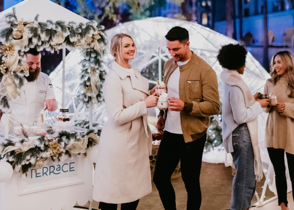 A chef behind a pop up drink station decorated white and evergreen garland. Couple in the forefront laughing with drinks. Two gals to the left chatting.