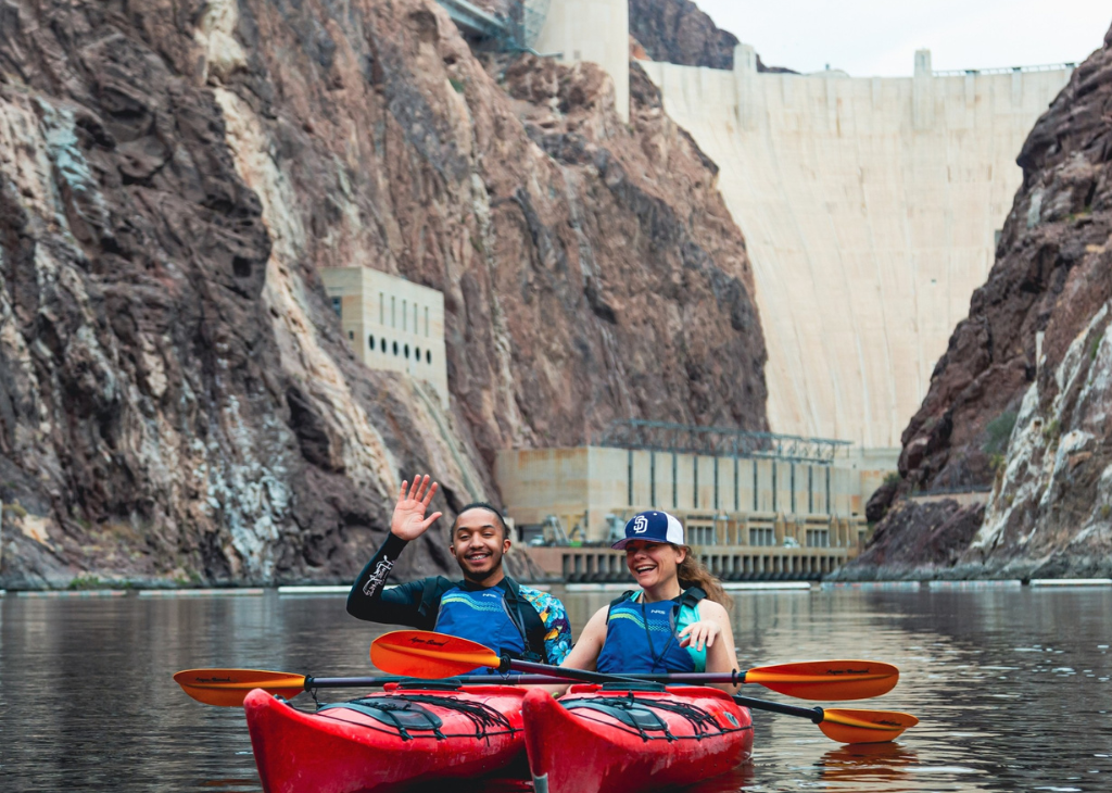 A man and a woman in red kayaks waving as they float on the Colorado River for a photo opportunity with the Hoover Dam as a backdrop.