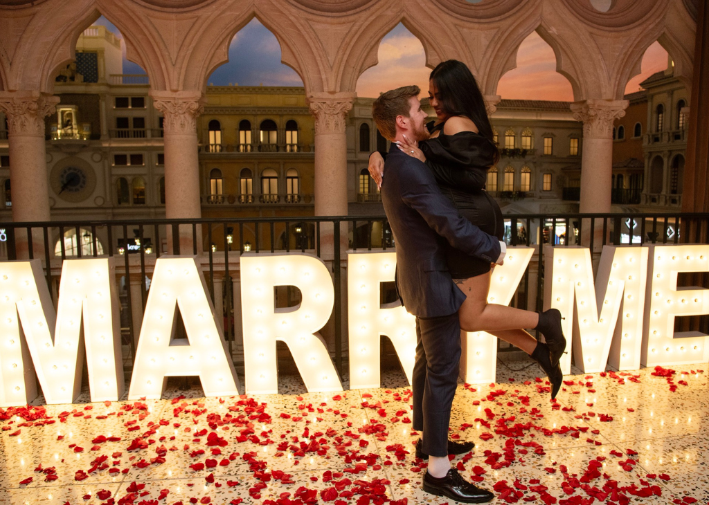 A couple where the female is jumping up in the arms of the man that just asked her to Marry Me with a giant sign in lights and rose pedals on the floor.